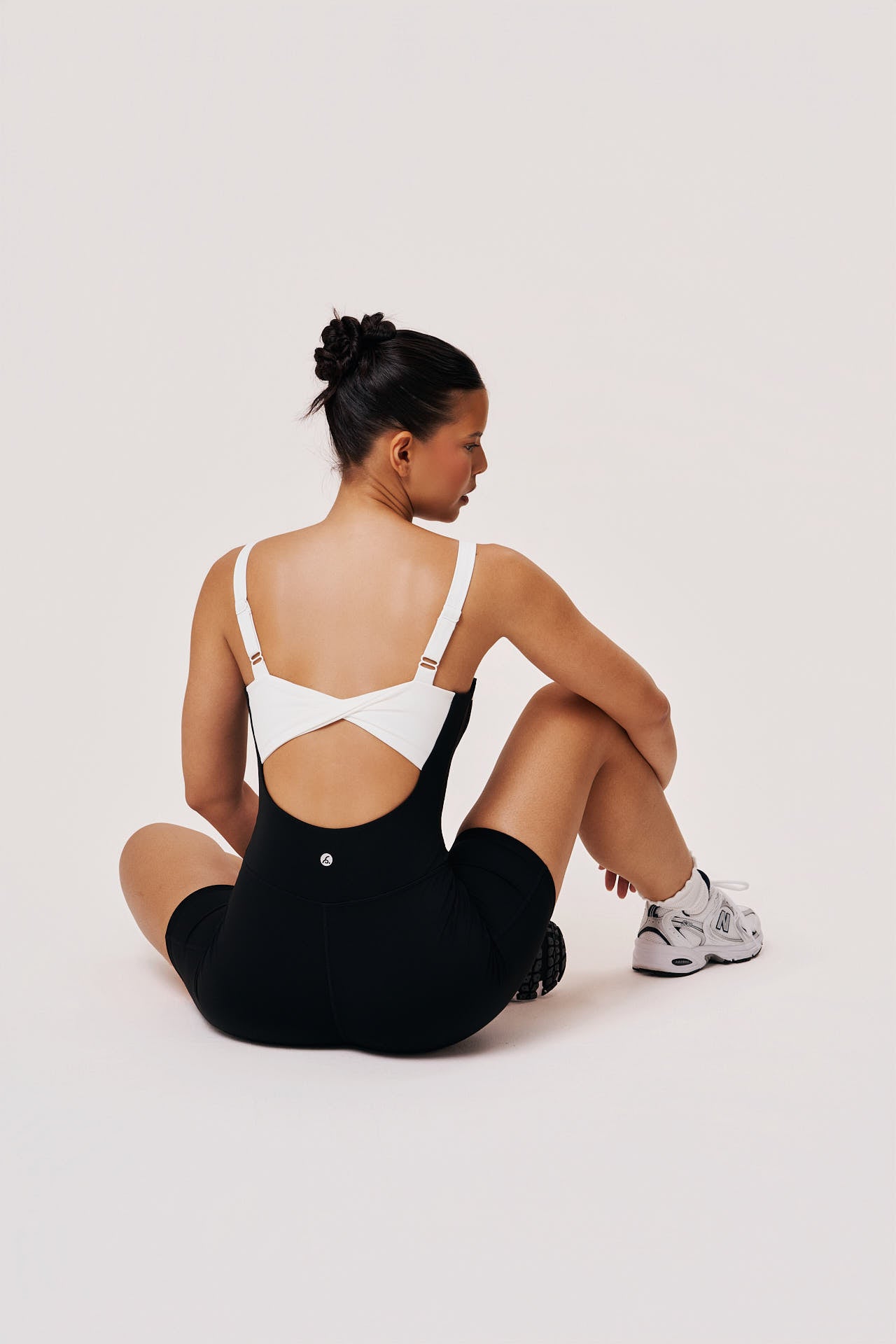 Woman sitting on a white floor wearing a black outfit with cut-out design and white sneakers.